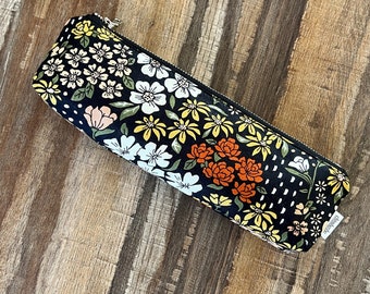 May include: Black pencil case with a floral pattern featuring yellow, orange, white, and green flowers on a black background. The zipper pull has a gold metal charm.
