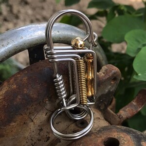 Puede incluir: Un llavero de metal plateado y dorado con un resorte y un anillo. El llavero está hecho de alambre y tiene un diseño único.