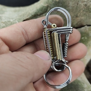 Könnte beinhalten: Ein silberner und goldener Schlüsselanhänger mit mehreren Schlaufen und einer Feder. Der Schlüsselanhänger ist aus Metall und hat eine polierte Oberfläche.