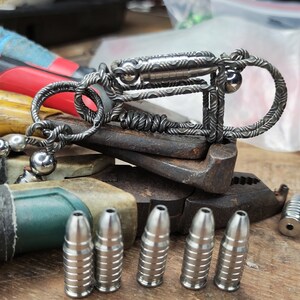 Może przedstawiać: Srebrny metalowy brelok do kluczy z plecionym wzorem i małym cylindrem. Brelok leży na szczypcach. Na pierwszym planie znajduje się sześć srebrnych metalowych przedmiot&oacute;w w kształcie pocisku.