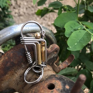 May include: A silver and gold metal key chain with a spring and a round ring. The key chain is attached to a rusty metal object.