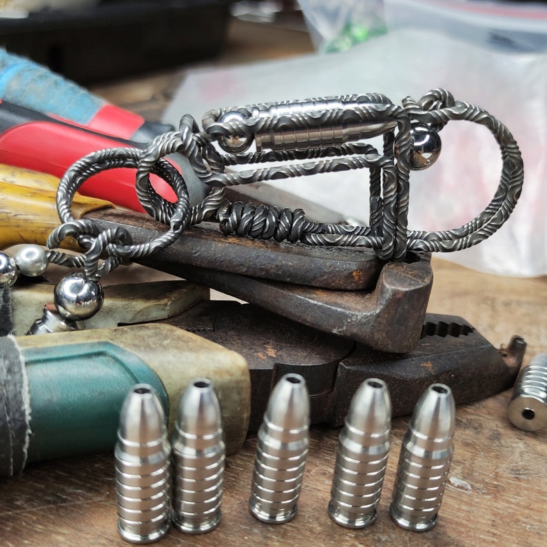 Może przedstawiać: Srebrny metalowy brelok do kluczy z plecionym wzorem i małym cylindrem. Brelok leży na szczypcach. Na pierwszym planie znajduje się sześć srebrnych metalowych przedmiot&oacute;w w kształcie pocisku.