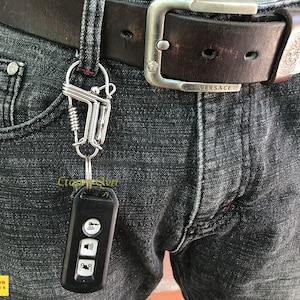 May include: A silver key chain with a spring-loaded clip attached to a black car key fob with three buttons. The key chain is attached to a belt loop on a pair of dark grey jeans.