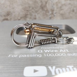 May include: A silver and gold metal keychain with a spring and a small ball. The keychain is presented to Q Wire Art for passing 100,000 subscribers on YouTube.