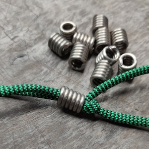 May include: A close-up of a green and black paracord bracelet with a silver metal bead. The bead is made of multiple coils of metal. There are several other silver metal beads scattered around the bracelet.