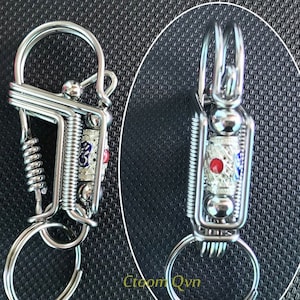 Puede incluir: Un llavero de metal plateado con un resorte y un pequeño amuleto con un diseño rojo y azul. El amuleto tiene un fondo plateado con un punto rojo en el centro y detalles azules y blancos alrededor del punto.