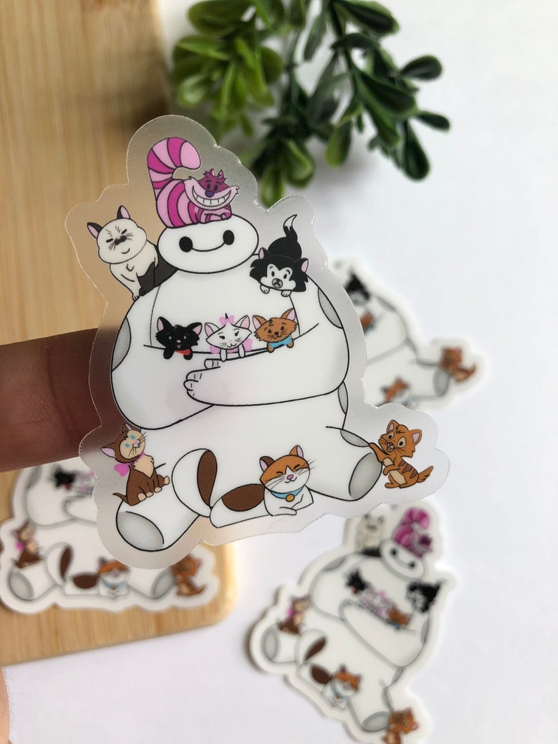 K&ouml;nnte beinhalten: Ein transparenter Aufkleber mit einem wei&szlig;en Robotercharakter mit einem l&auml;chelnden Gesicht, der mehrere Comic-Katzen h&auml;lt. Die Katzen sind in verschiedenen Farben und haben unterschiedliche Gesichtsausdr&uuml;cke. Der Aufkleber befindet sich auf einer Holzoberfl&auml;che.