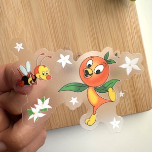 May include: A clear sticker with an orange cartoon bird with a yellow beak and orange feet. The bird is holding a green leaf in its right wing. There is a bee with black and yellow stripes and white wings flying next to the bird. There are white flowers scattered around the bird and bee.
