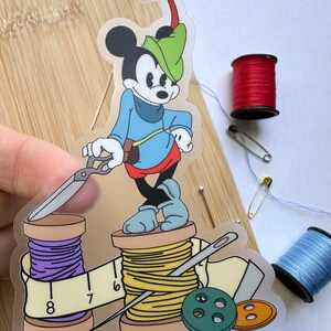 May include: A clear sticker of Mickey Mouse as Robin Hood, holding scissors, standing on thread spools. Includes a measuring tape, buttons, and needles. Additional spools of red and blue thread, and a safety pin are also visible.