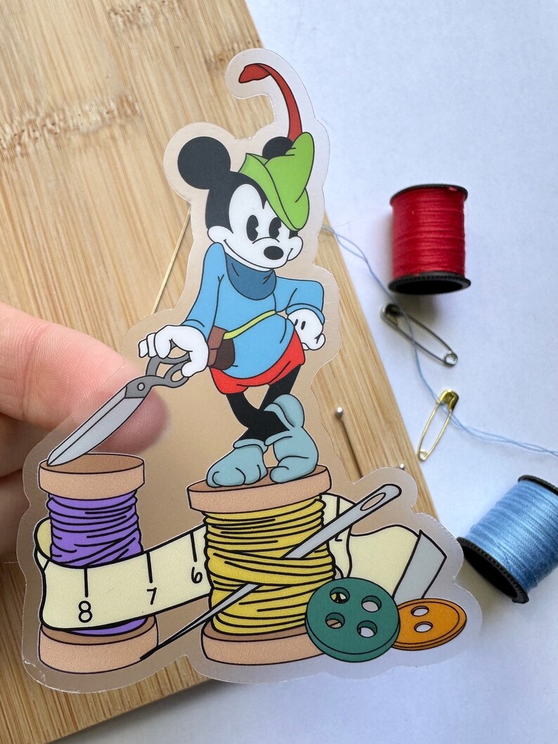 May include: A clear sticker of Mickey Mouse as Robin Hood, holding scissors, standing on thread spools. Includes a measuring tape, buttons, and needles. Additional spools of red and blue thread, and a safety pin are also visible.
