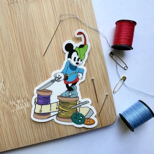 May include: A sticker of a cartoon mouse in a blue outfit and green hat, standing on spools of thread, scissors, and a measuring tape. Red and blue spools of thread, needles, and a safety pin are on a wooden cutting board.