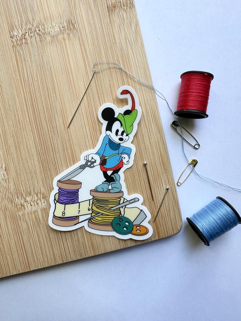 May include: A sticker of a cartoon mouse in a blue outfit and green hat, standing on spools of thread, scissors, and a measuring tape. Red and blue spools of thread, needles, and a safety pin are on a wooden cutting board.