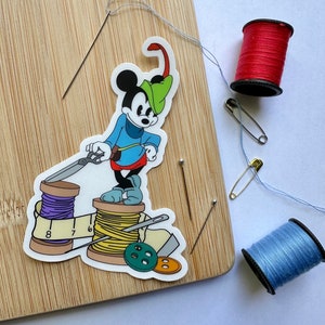 May include: A sewing-themed image with a Mickey Mouse sticker dressed as a tailor, alongside sewing needles, red and blue thread spools, safety pins, and buttons. The sticker is on a wooden cutting board.