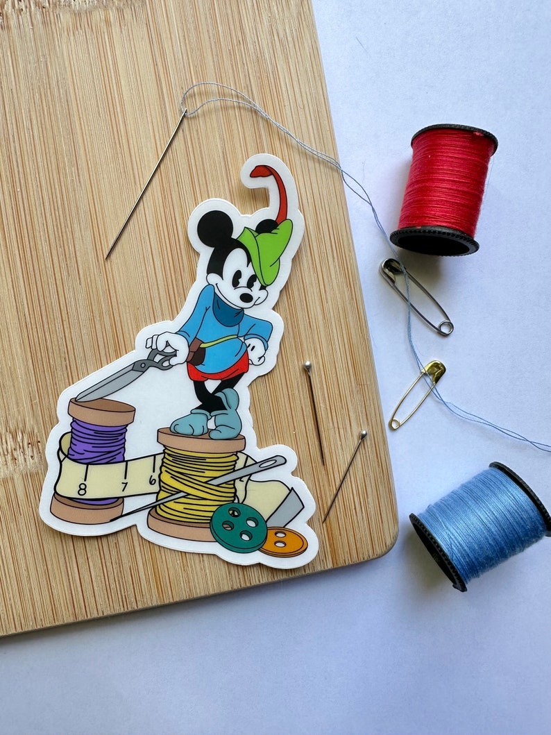 May include: A sewing-themed image with a Mickey Mouse sticker dressed as a tailor, alongside sewing needles, red and blue thread spools, safety pins, and buttons. The sticker is on a wooden cutting board.
