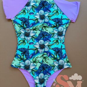 May include: One-piece swimsuit with a floral and butterfly pattern in shades of blue, teal, and purple. The swimsuit features short sleeves and trim in a solid lavender color. The design includes stylized flowers, butterflies, and crystal-like accents.