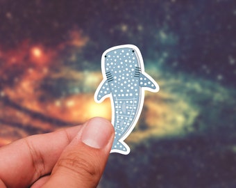 May include: A blue and white whale shark sticker with white spots. The sticker is held in a hand against a blurred background of a galaxy.
