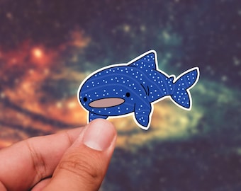 May include: A blue whale shark sticker with white polka dots and a white outline. The sticker has a cartoon style with a smiling face. The background is a blurred image of a colorful sky.