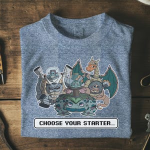 Puede incluir: Camiseta gris con un gráfico colorido de personajes de dibujos animados con trajes espaciales, incluyendo un dragón, una tortuga y un gato. El texto "CHOOSE YOUR STARTER...." está impreso en un rectángulo blanco en la parte inferior de la camiseta.