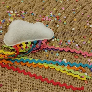 May include: A whimsical felt cloud with colorful, wavy ribbons in red, orange, yellow, green, and blue. The cloud is white, and the ribbons cascade downward. Confetti in various colors surrounds the cloud on a textured, brown surface.
