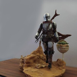 May include: A silver and brown Mandalorian action figure with a green alien child in a floating pod. The figure is standing on a brown rock base.
