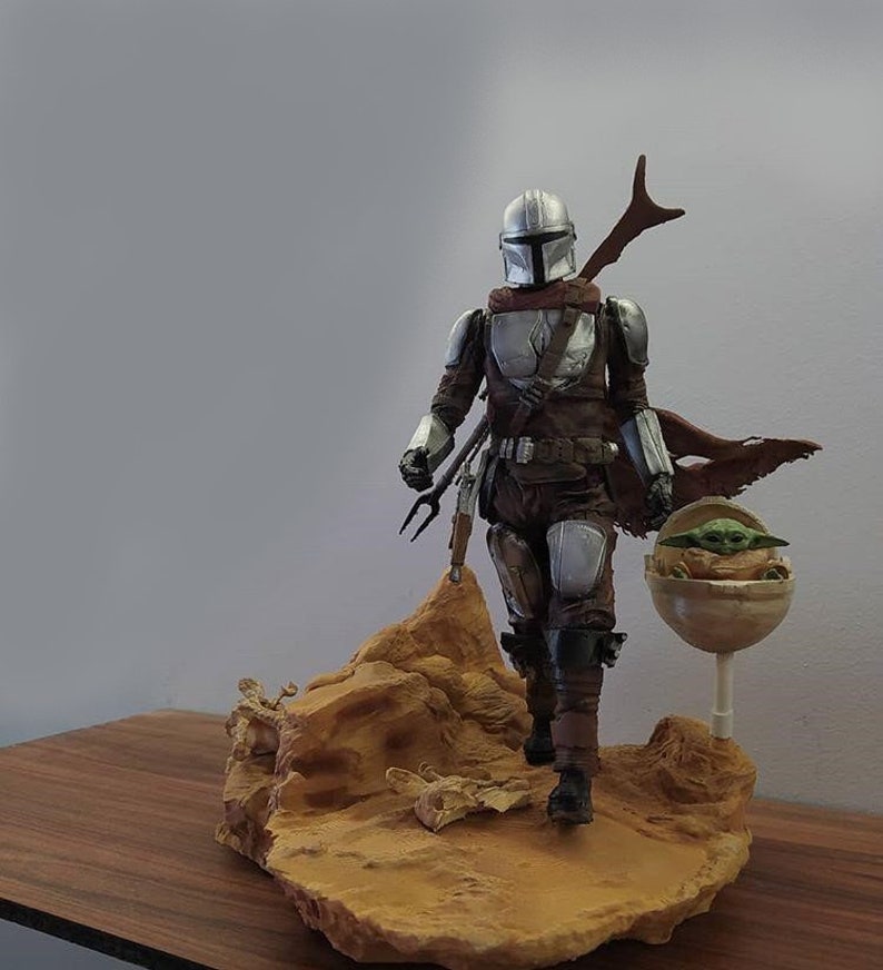 May include: A silver and brown Mandalorian action figure with a green alien child in a floating pod. The figure is standing on a brown rock base.