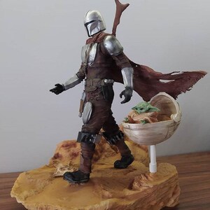 May include: A silver and brown Mandalorian action figure with a removable cape and a green baby Yoda action figure in a floating pod. The figures are standing on a brown desert-themed base.