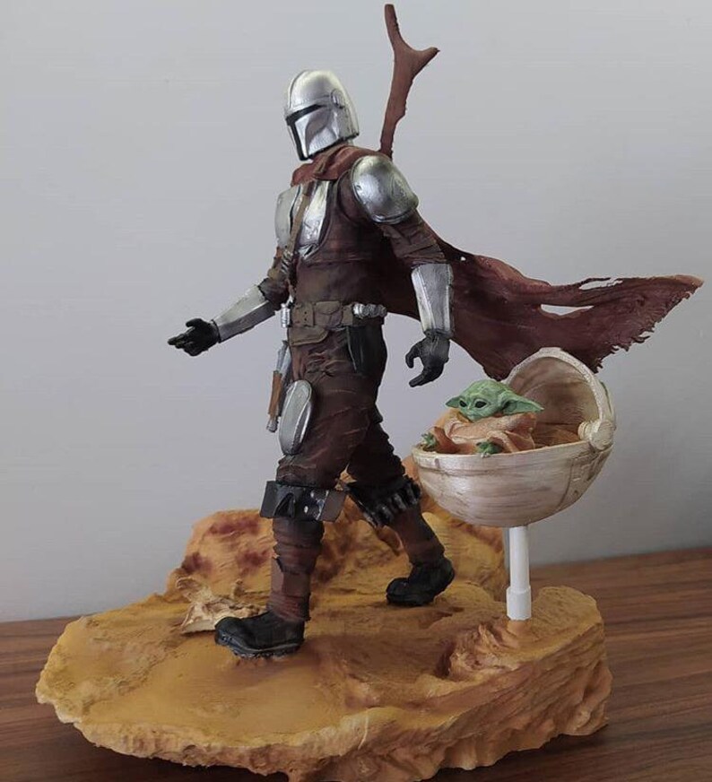 May include: A silver and brown Mandalorian action figure with a removable cape and a green baby Yoda action figure in a floating pod. The figures are standing on a brown desert-themed base.