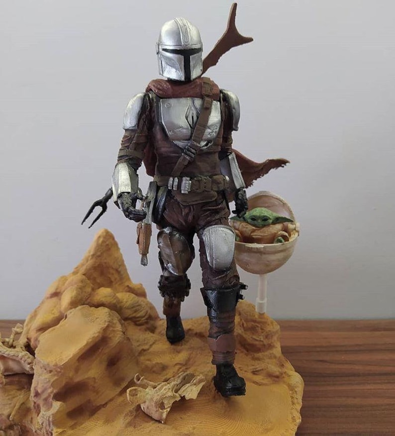 May include: A silver and brown action figure of a Mandalorian warrior holding a small green alien in a floating pod. The figure is standing on a rocky base.