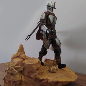 May include: A silver and brown action figure of a character wearing a helmet and holding a weapon, standing on a rocky base.