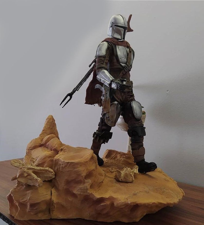 May include: A silver and brown action figure of a character wearing a helmet and holding a weapon, standing on a rocky base.
