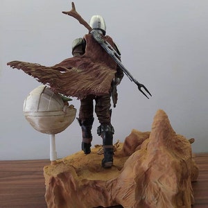 May include: A brown and gray action figure of a character from the Star Wars series, The Mandalorian, standing on a desert-themed base. The figure is holding a weapon and has a cape flowing behind it. A small, round, white object is on the base next to the figure.