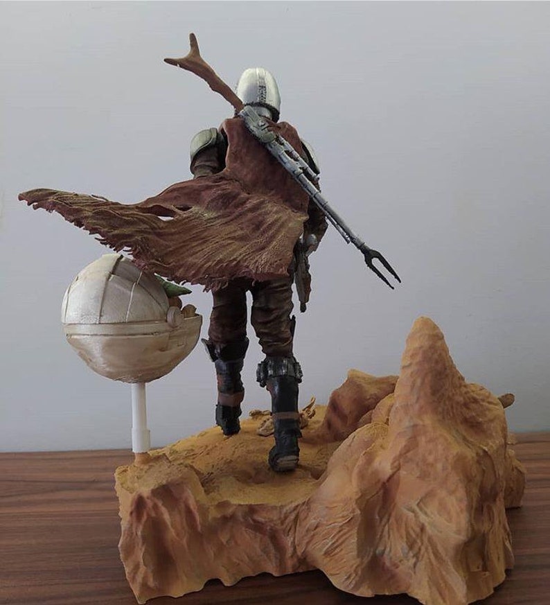 May include: A brown and gray action figure of a character from the Star Wars series, The Mandalorian, standing on a desert-themed base. The figure is holding a weapon and has a cape flowing behind it. A small, round, white object is on the base next to the figure.