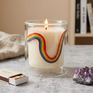 May include: A lit, white candle in a clear glass holder. A rainbow design curves around the candle. A matchbox with the word "DESIGNER" is in the foreground. An amethyst geode is to the right.
