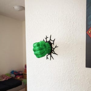 Könnte beinhalten: Eine grüne Faust, die durch eine weiße Wand bricht, mit einem schwarzen Rissdesign darum. Die Faust hat eine leuchtend grüne Farbe, und die Wand ist strukturiert. Das Design erinnert an die Kraft eines Superhelden.