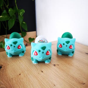 Könnte beinhalten: Drei grüne und blaue Plastikfiguren eines Pokemon-Charakters, Bisasam, die ein weißes Kinder-Überraschungsei halten.