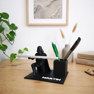 Könnte beinhalten: Ein schwarzer Schreibtisch-Organizer im geometrischen Stil mit einer knienden Figur, die Darth Vader ähnelt. Die Figur hält einen weißen Stift, während ein angebrachter quadratischer Stiftehalter einen grünen und einen schwarzen Stift enthält. Die Basis trägt den Namen MARTIN.