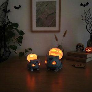 Könnte beinhalten: Zwei graue, Bulbasaur-förmige Lampen mit leuchtend orangefarbenen Augen und Mündern, die jeweils einen beleuchteten Kürbis auf dem Rücken tragen. Die Lampen strahlen ein warmes Licht aus und betonen die detaillierten Designs der Kreaturen.