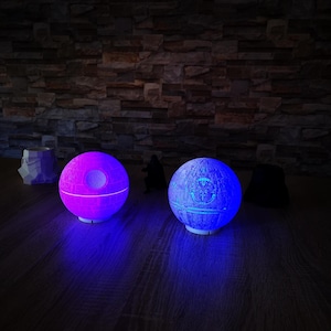 Könnte beinhalten: Zwei kugelförmige Lampen, eine lila und die andere blau leuchtend, die dem Todesstern aus Star Wars ähneln. Die Lampen stehen auf einer Holzoberfläche, mit einer Backsteinmauer im Hintergrund. Zusätzliche Figuren zum Thema Star Wars sind ebenfalls vorhanden.