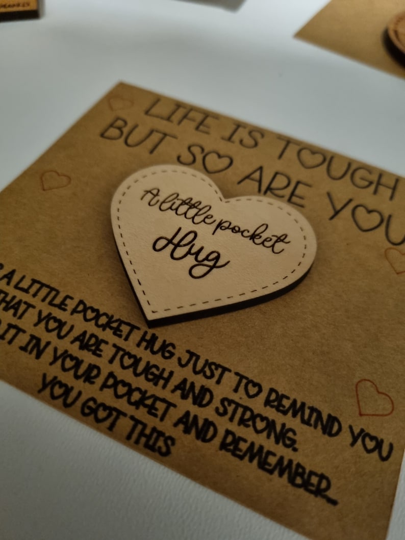 以下が含まれることがあります： 「LIFE IS TOUGH BUT SO ARE YOU」の文字が入った薄茶色のカードと、「A little pocket Hug」と書かれたハート型の木片。カードには「A LITTLE POCKET HUG JUST TO REMIND YOU THAT YOU ARE TOUGH AND STRONG. IT IN YOUR POCKET AND REMEMBER... YOU GOT THIS.」の文字も入っています。