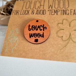 May include: A round wooden charm with the text "touch wood" and a small four-leaf clover. The charm is on a brown card with the text "Touch Wood for Luck & Avoid Tempting Fate".