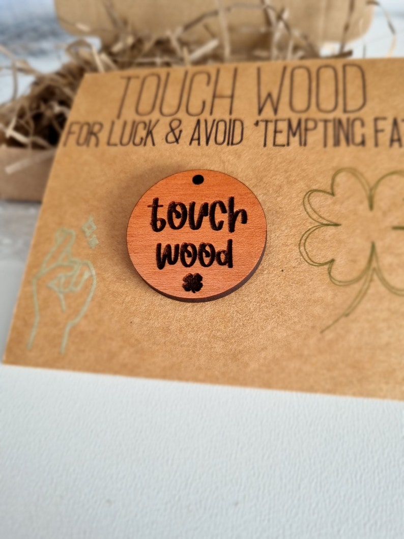May include: A round wooden charm with the text "touch wood" and a small four-leaf clover. The charm is on a brown card with the text "Touch Wood for Luck & Avoid Tempting Fate".