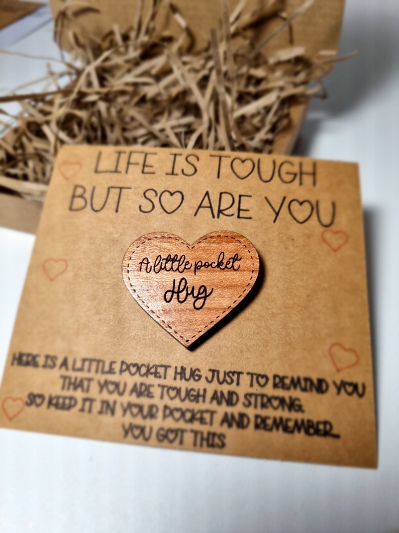 以下が含まれることがあります： 「Life is tough but so are you」の文字と「A little pocket hug」の木製ハートが描かれた茶色のカード。カードには励ましのメッセージが書かれています。背景には、細かく刻まれた紙が入った箱があります。