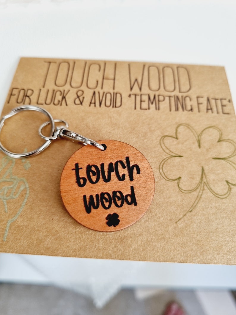 May include: A wooden keychain with a round tag that says "touch wood" in black lettering. The keychain is attached to a silver key ring. The tag also has a small four-leaf clover carved into it.