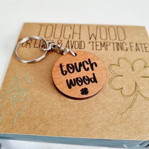 May include: A wooden keychain with a round charm that says "touch wood" in black lettering. The charm is attached to a silver key ring. The background is a brown paper card with the text "Touch Wood for Luck & Avoid Tempting Fate" in black lettering.