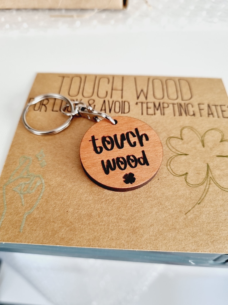 May include: A wooden keychain with a round charm that says "touch wood" in black lettering. The charm is attached to a silver key ring. The background is a brown paper card with the text "Touch Wood for Luck & Avoid Tempting Fate" in black lettering.