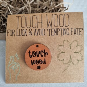 May include: A brown wooden circle with the text "touch wood" and a small four-leaf clover engraved on it. The circle is on a brown card with the text "TOUCH WOOD FOR LUCK & AVOID 'TEMPTING FATE'".