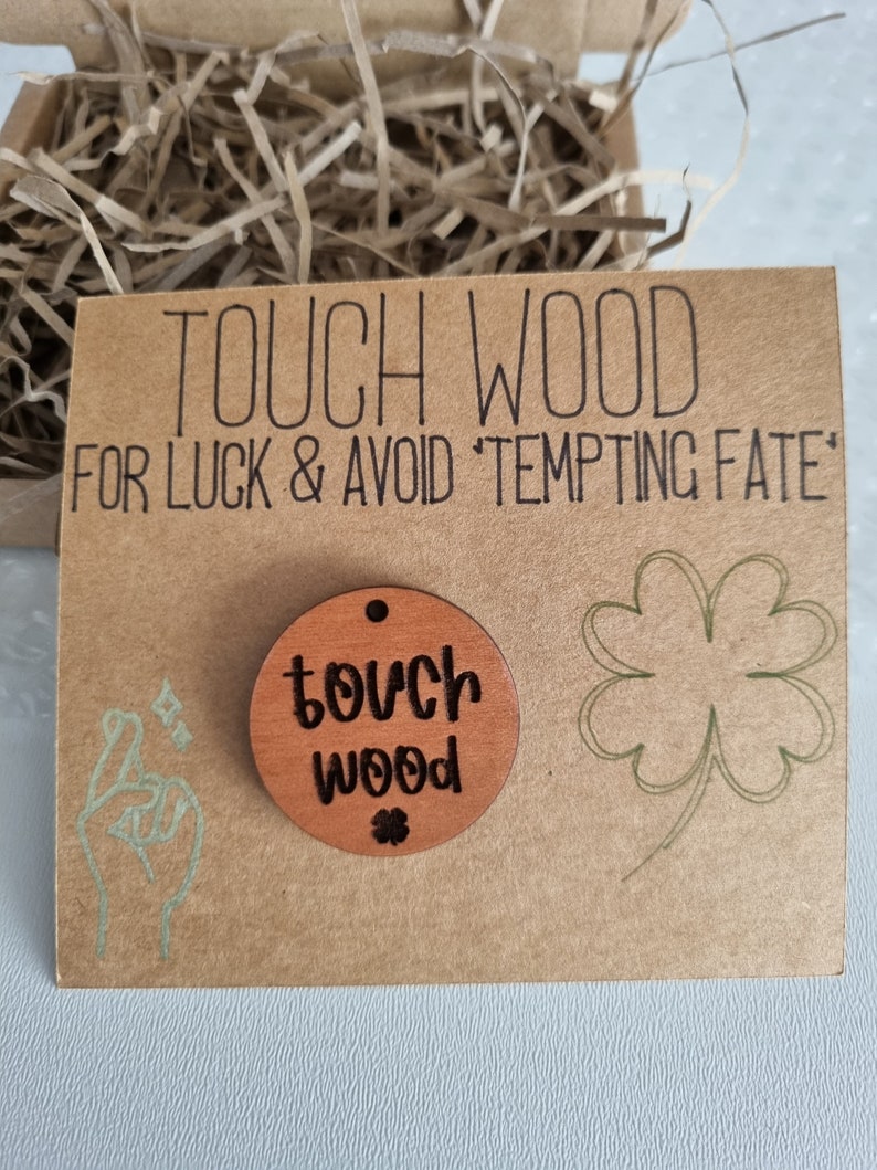 May include: A brown wooden circle with the text "touch wood" and a small four-leaf clover engraved on it. The circle is on a brown card with the text "TOUCH WOOD FOR LUCK & AVOID 'TEMPTING FATE'".
