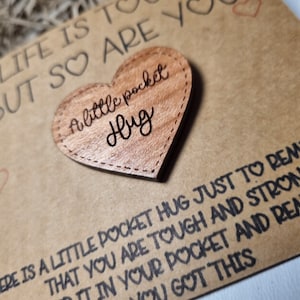 以下が含まれることがあります： 「a little pocket hug」と刻まれた木製のハート型のポケットハグ。ハートは、「Life is tough but so are you」というテキストと他の応援メッセージが書かれた茶色のカードの上に置かれています。