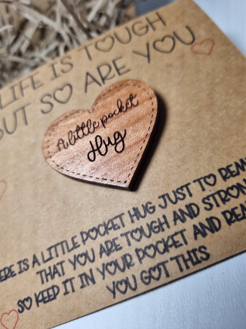 以下が含まれることがあります： 「a little pocket hug」と刻まれた木製のハート型のポケットハグ。ハートは、「Life is tough but so are you」というテキストと他の応援メッセージが書かれた茶色のカードの上に置かれています。