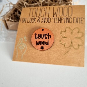 May include: A round wooden charm with the text "touch wood" and a four-leaf clover symbol. The charm is on a brown card with the text "Touch Wood for Luck & Avoid 'Tempting Fate'".
